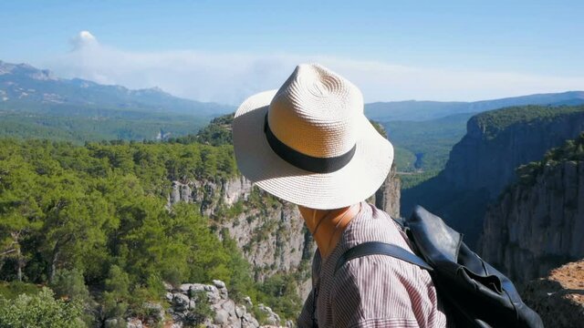 A young woman hiker has reached the mountain peak and enjoys a beautiful view of the forest and canyon.Young attractive woman traveler stands on the top of the mountain and looks into the distance at