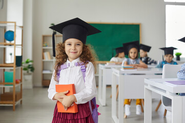Portrait of a beautiful little curly elementary school graduate girl wearing an academic hat. Child...