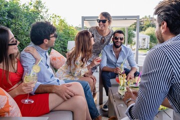 A group of laughing young people gathers at the aperitif party on the terrace. Millennials congregated in the garden joke together. - obrazy, fototapety, plakaty