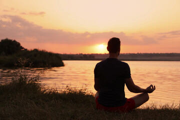 Man meditating near river at sunset, back view. Space for text