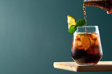 Cuba Libre or Long Island Cocktail with pouring cola, rum and lime in glass on wooden board ob dark blue background. Refreshing summer drink