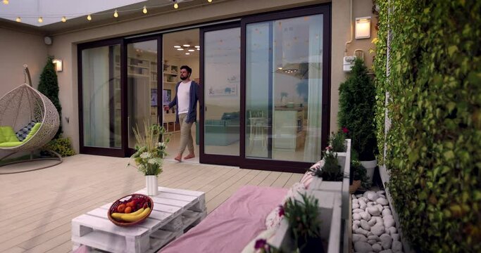 man opens wooden sliding doors, going out on rooftop patio with cozy lounge zone