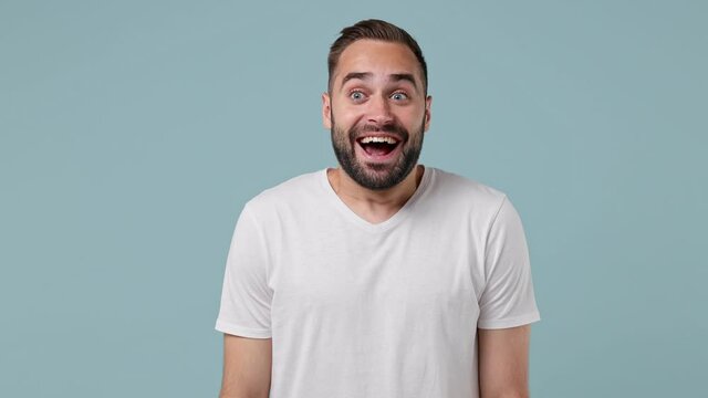 Smiling amazed surprised fun exultant young brunet man 20s wears white t-shirt keeping mouth wide open looking aside say yes isolated on pastel light blue background. People emotions lifestyle concept