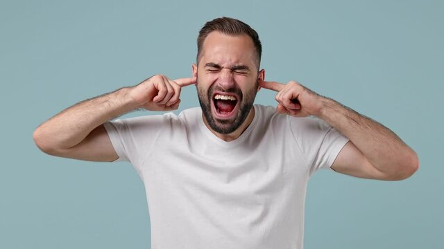 Displeased irritated sad angry young brunet man 20s wears white t-shirt closed eyes cover ears do not want to listen scream isolated on pastel light blue background. People emotions lifestyle concept