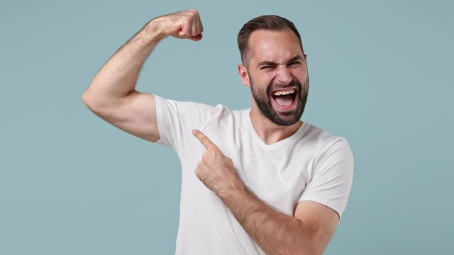 Strong sporty fitness young brunet man 20s wears white t-shirt showing biceps muscles on hand demonstrating strength power isolated on pastel light blue background. People emotions lifestyle concept