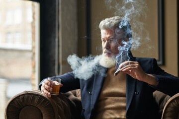 Elegant mature businessman smoking cigar with fume and holding a glass of whisky while relaxing in...
