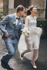 Stylish happy bride and groom running on background of old church in rainy street. Provence...