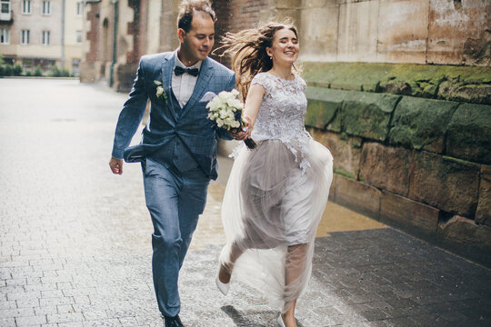 Beautiful emotional wedding couple smiling and having fun in rain in european city. Provence wedding. Stylish happy bride and groom running on background of old church in rainy street.