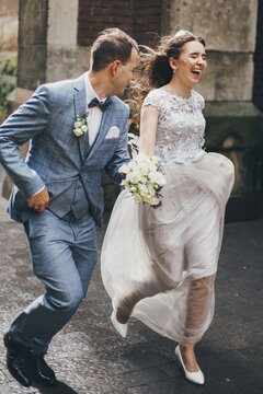 Stylish happy bride and groom running on background of old church in rainy street. Provence wedding. Beautiful emotional wedding couple smiling and having fun in rain in european city.