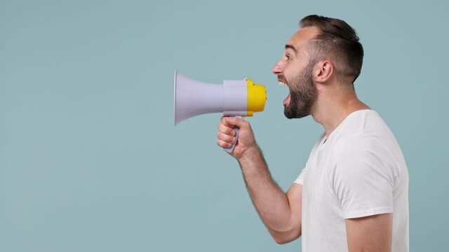 Side view profile shocked excited astonished promoter young bearded brunet man 20s wears white t-shirt scream in megaphone announces discounts sale hurry up isolated on pastel light blue background