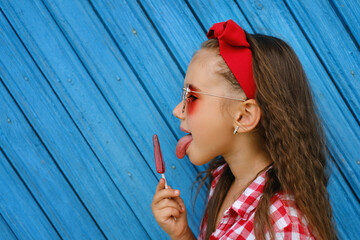 A girl in a checkered red shirt and sunglasses holds a lollipop in her hands, licking it with her tongue. A child with a candy on a blue board background  - obrazy, fototapety, plakaty
