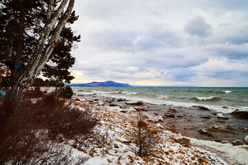 Nature landscape with dramatic sky and clouds, lake with ice, shore with snow and dark trees in...
