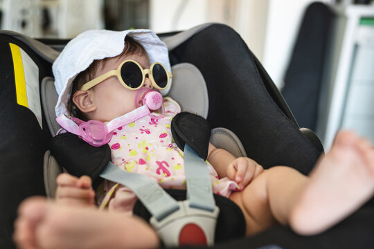 Small Caucasian Baby Four Months Old Sitting In The Car Seat At Home In Room With Eyeglasses And Hat On Her Head Ready For Travel To Go Out On Vacation