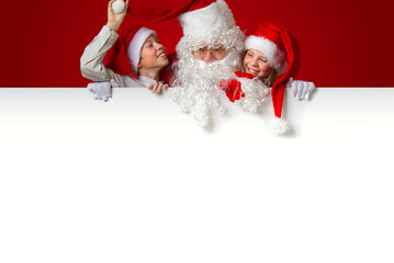 satna claus with funny children in red christmas gnome hats peeking out from behind a blank white banner, mockup and copy space