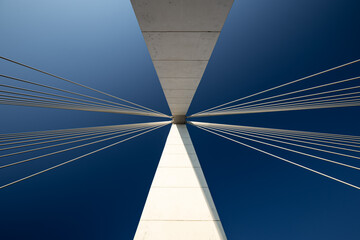 Fototapeta premium Minimalist abstract architecture shot featuring a white concrete pillar of a suspension bridge with two bunches of suspension cables against a clear blue sky