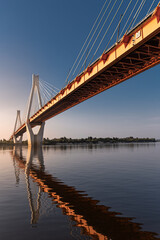 A view from the ground to a suspension bridge with two huge pillars reflecting in a river at sunrise in a rural area