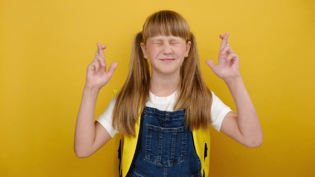 Portrait cute blonde kid schoolgirl keep fingers crossed, wears backpack posing isolated over yellow color background wall in studio. People childhood lifestyle concept wait for special moment 