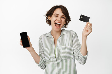 Happy laughing girl showing credit card and mobile phone screen, dancing with smartphone and laughing, shopping online in app, standing over white background