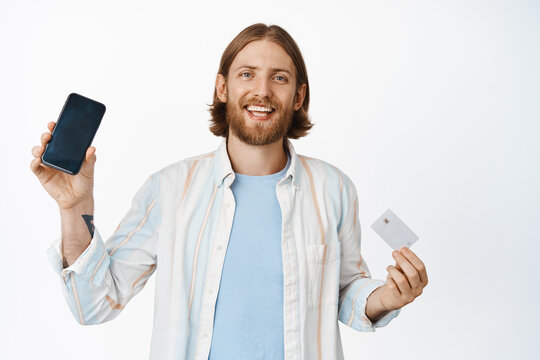 Image of handsome bearded man showing smartphone screen, app interface and credit card discount, smiling pleased, advertising application, standing against white background