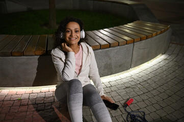 Top view of a female athlete with headphones smiling at camera sitting on the grounds and leaning on illuminated stone bench