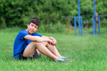 teenage boy exercising outdoors, sports ground in the yard, he sits on the green grass of the playground, healthy lifestyle