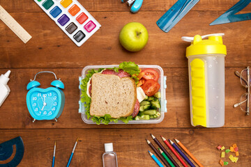 School lunch box with sandwich, vegetables, water, fruit on a wooden background. School supplies,...