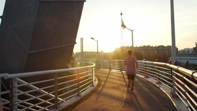 a guy in a T-shirt and shorts is runing across the bridge at dawn or sunset. jogging on the background of the urban landscape. the silhouette of a runner moving away from the camera.