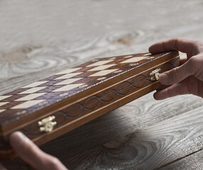 Close up of a carved wooden chess box in hands. Hands are going to close the folding chessboard. Wooden chess in a box on a textured table with a real forest. Chess board with lock
