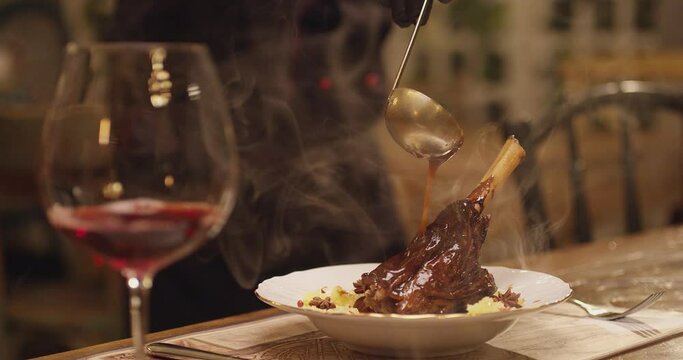 Spoon pouring sauce on meat. Cooked lamb knuckle . Dark red sauce poured on meat of lamb in plate with salad . Chef's Table with glass of wine and cooking show inspired footage .  Slow motion .