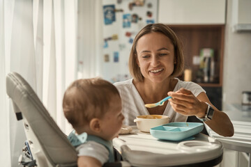 Obraz premium Baby sits at a children's little table. Mum feeds the child with porridge. Mother gives baby food from a spoon. High quality 4k footage