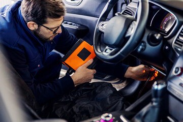 Car diagnostics at technical inspection, car electronics. A man in a blue uniform holds a digitizing device and checks the condition of the string inside the car under the steering wheel