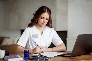 Beautiful nurse in a white medical suit sits at a table in front of a laptop and makes notes.
