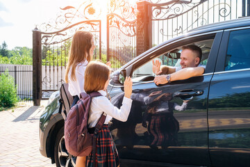 Smiling middle-aged father in the car, leaving her daughters and sisters in school uniform at school and gesturing goodbye.