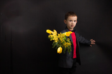 a boy with vitiligo in a red T-shirt, gray jacket and bow tie, with a bouquet of mimosa and yellow tulips, on a black studio background
