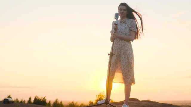 beautiful woman in white dress on top of mountain, on edge of abyss and sings song into shiny retro vintage microphone. Attractive lady singer on background of sunset or dawn sky. Sun shine in camera.