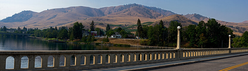 Panoramic landscape view of a bridge over a finger of Lake Chelan at Chelan, Washington with the arid hills in the background and greenery in the foreground in a horizontal format..