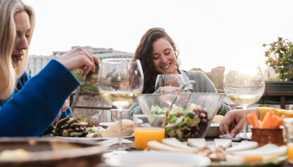 Happy diverse people eating vegan food with wine outdoors in sunset at patio restaurant - Focus on center woman face