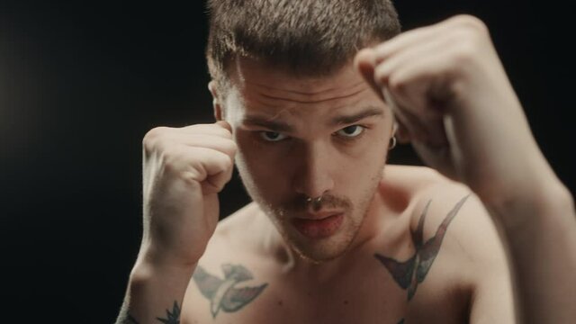 Fashion portrait of brutal naked man in tattoos, having earring in his nose and ears, ready for punches, strong muscle guy posing in dark studio, looking in camera with serious view. 