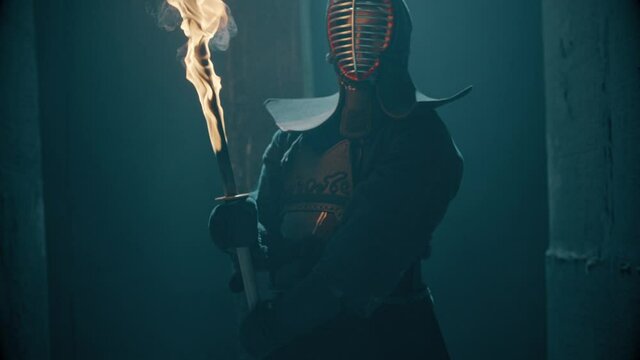 Portrait of a Japanese kendo fighter hold a burning sword . Epic Kendo warrior stands in armor in the fog and looking at the camera with his fire shinai . Mysterious video scene . Shot on Arri Alexa .