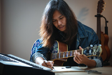 Asian musician who have long hair and wearing jean jacket playing guitar and writing a song in...