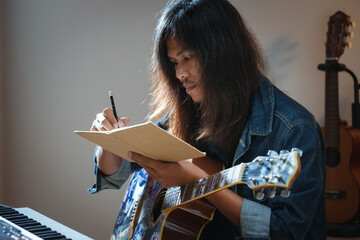 Asian musician who have long hair and wearing jean jacket playing guitar and writing a song in...
