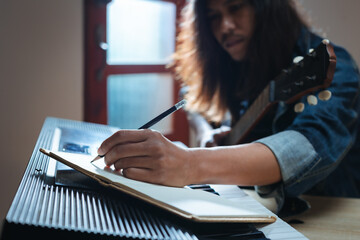 Asian musician who have long hair and wearing jean jacket playing keyboard and guitar and writing a song in notebooks in the dark room and lighting.
