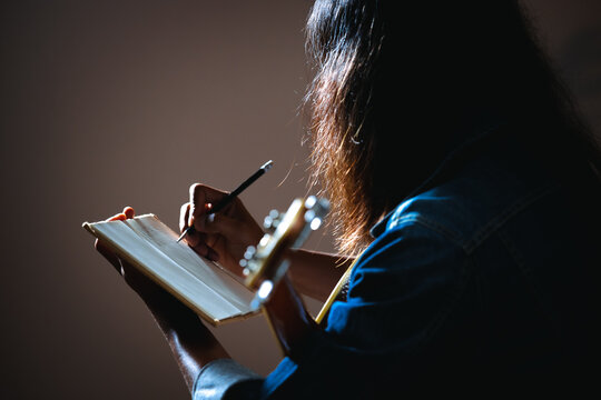 Asian musician who have long hair and wearing jean jacket playing guitar and writing a song in notebooks in the dark room and lighting.