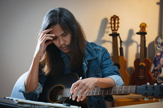 Asian musician who have long hair and wear jean jacket is playing guitar and so stressed to think about lyric and write a song in notebooks in the dark room.