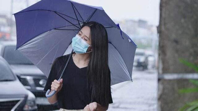 young asian Woman wear protective face mask holding blue umbrella standing on the street side walk, shower rainy season, pouring rain, risk of getting sick, hard pouring rain, reach out hand curious