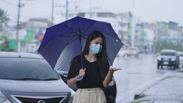 Young asian black hair woman walks outside among a heavy rain caused by tropical typhoon, casual girl feels sick of getting wet, holding umbrella walk on city street footpath with car parking behind
