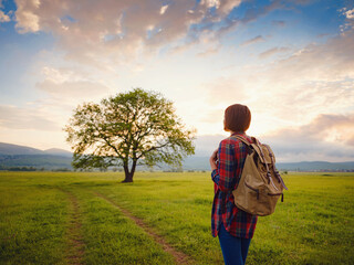 Asian traveler woman walk on the dirty road with sunshine and oak tree. In a plaid shirt and with a backpack. The end and the idea of adventure, travel, and discovery