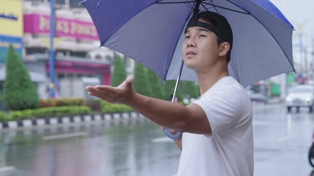 Young asian man holding a blue umbrella standing on the street sidewalk, shower rainy season, pouring rain, risk of getting sick, sick of stuck in rain, reach out hand curious, tropical typhoon comin 