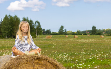 Naklejka na ściany i meble Happy girl sitting on a haystack in the middle of a green meadow