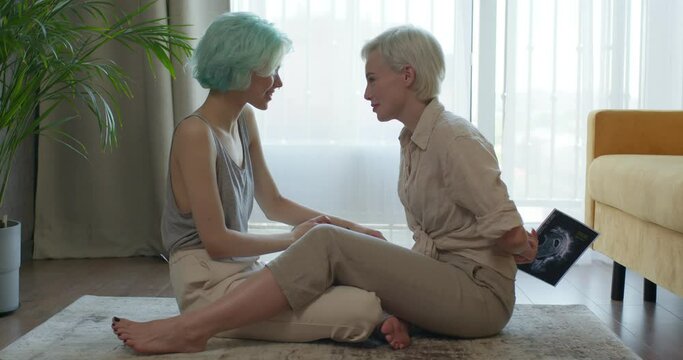 Caucasian Lesbian Couple Have Fun, Getting News ABout Pregnancy, Future Mothers, Side View On Happy Ladies In Casual Clothes Sit On Floor At Home, Laughing, Talking, Hugging, Celebrating Pregnancy